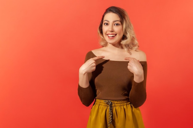 Portrait d'une charmante femme heureuse en blouse décontractée et pantalon souriant et pointant vers elle-même, fière de son propre succès, se vantant de ses réalisations. Tourné en studio intérieur isolé sur fond rouge