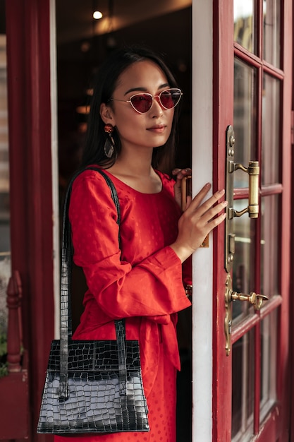 Portrait d'une charmante femme séduisante en robe rouge, des lunettes de soleil élégantes ouvrent une porte en bois et tiennent un sac à main noir