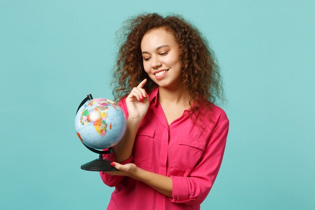 Portrait d'une charmante fille africaine en vêtements décontractés tenant dans les mains un globe terrestre isolé sur fond bleu turquoise en studio. Les gens émotions sincères, concept de style de vie. Maquette de l'espace de copie.