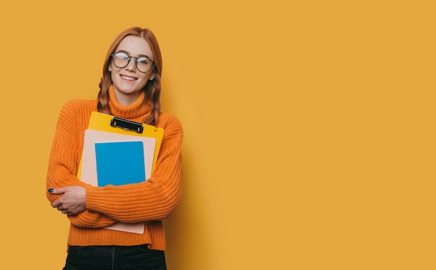 Portrait d'une charmante jeune étudiante avec des taches de rousseur et des cheveux rouges portant des lunettes de soleil regardant la caméra en souriant tout en tenant ses livres isolés sur jaune.