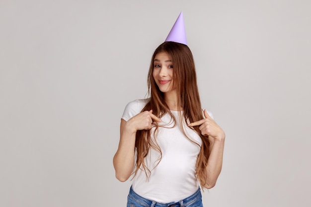 Portrait d'une charmante jeune femme joyeuse dans un cône de fête sur la tête pointant vers elle-même célébrant son anniversaire portant un t-shirt blanc Studio d'intérieur tourné isolé sur fond gris