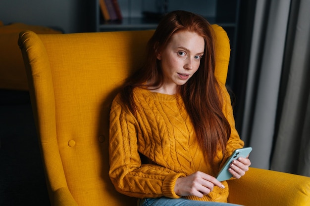 Portrait d'une charmante jeune femme rousse utilisant un smartphone assis dans un fauteuil jaune souriant regardant la caméra