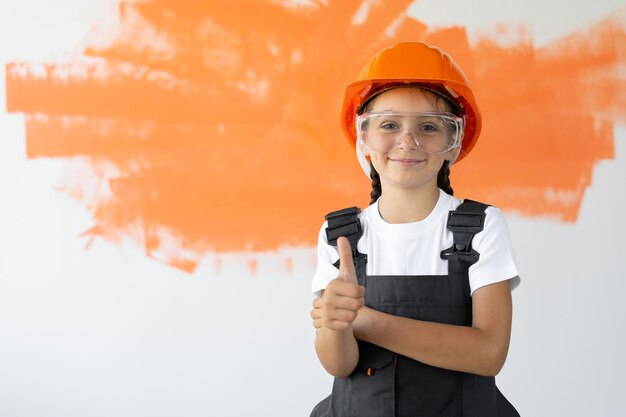 Portrait d'une charmante jeune fille dans un casque orange sur la tête. leva le pouce en regardant le photographe. Un sourire sur votre visage. les lunettes sont portées sur le visage.