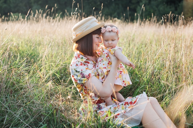 Photo portrait d'une charmante mère paisible, assise sur l'herbe dans le champ en plein air et serrant sa petite fille mignonne dans ses bras. mère et fille s'amusant à l'extérieur