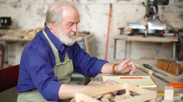 Portrait d'un charpentier aux cheveux gris à une table dans l'atelier avec divers outils pour le travail manuel