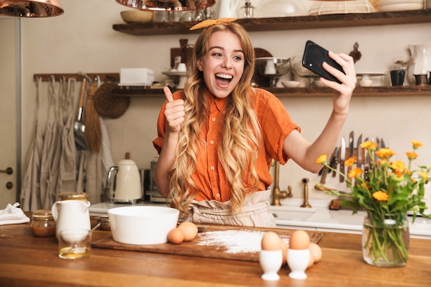 Portrait d'un chef heureux jeune fille blonde émotionnelle cuisine à la cuisine prendre un selfie par téléphone portable montrant le geste du pouce vers le haut.