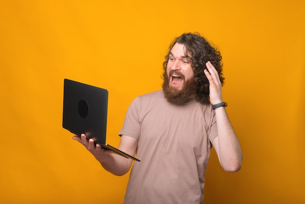 Portrait de choqué jeune homme barbu aux cheveux longs à la surprise de l'ordinateur portable sur jaune