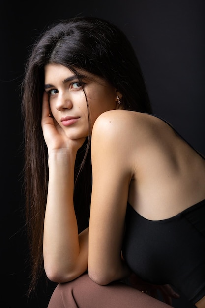 Portrait classique en studio d'une jeune brune vêtue d'un haut noir assise sur une chaise sur fond noir