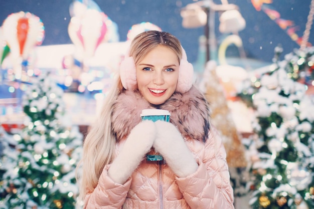 Portrait Close-Up of Beautiful Happy Female Model Looking Camera Holding Coffee Cup in White Mitaines Rose Hiver Cache-oreilles Snowy New Year Tree et Christmas Fair Attraction