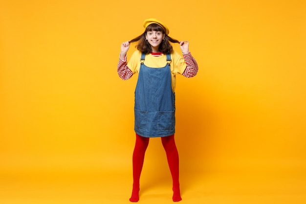 Portrait complet d'une adolescente drôle en béret français et robe d'été en jean tenant les cheveux isolés sur fond de mur jaune en studio. Concept de mode de vie des émotions sincères des gens. Maquette de l'espace de copie.