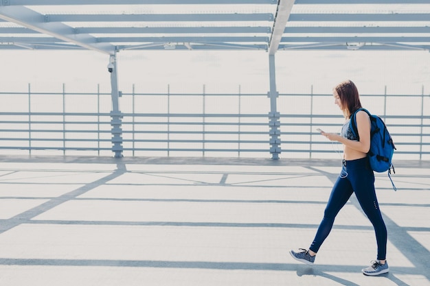 Le portrait complet d'une adolescente ou d'une jeune sportive active porte des leggings et des baskets haut de gamme porte un sac à dos apprécie une musique agréable dans la liste de lecture utilise un téléphone portable et des écouteurs modernes