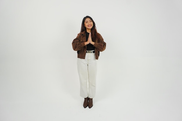 Un portrait complet du corps d'une jeune femme asiatique en tenue décontractée faisant un geste de salut avec les deux mains tout en souriant debout sur un fond blanc isolé