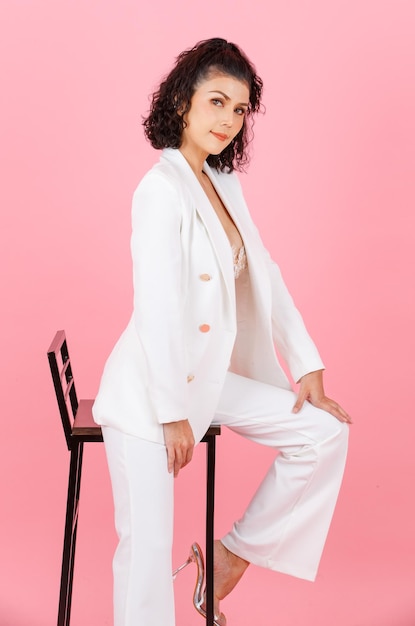 Portrait complet du corps tourné en studio d'une femme d'affaires prospère avec une coiffure frisée sexy asiatique en costume décontracté à la mode blanc avec un soutien-gorge en dentelle lingerie en dentelle assis sur une chaise haute posant sur fond rose
