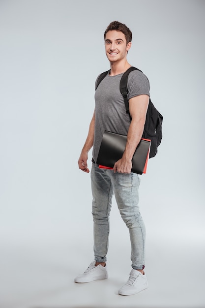 Portrait complet d'un étudiant souriant heureux debout avec sac à dos et dossiers sur blanc