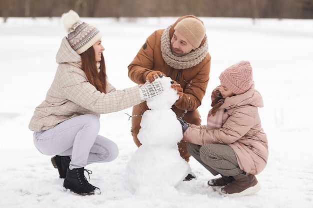 Portrait complet d'une famille heureuse construisant un bonhomme de neige ensemble tout en profitant d'une promenade à l'extérieur en hiver, espace pour copie