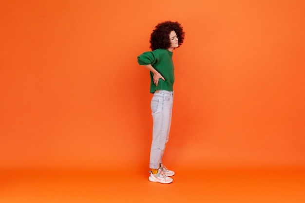 Portrait complet d'une femme avec une coiffure afro portant un chandail de style décontracté vert touchant la douleur dans le bas du dos ayant des problèmes de rein Prise de vue en studio intérieur isolée sur fond orange