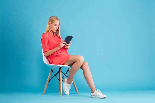 Portrait complet d'une femme décontractée concentrée en robe rouge à l'aide d'une tablette assise sur la chaise isolée sur le mur bleu