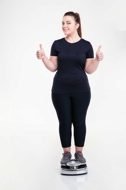 Portrait complet d'une femme épaisse et heureuse debout sur une balance et montrant les pouces vers le haut isolé sur un mur blanc