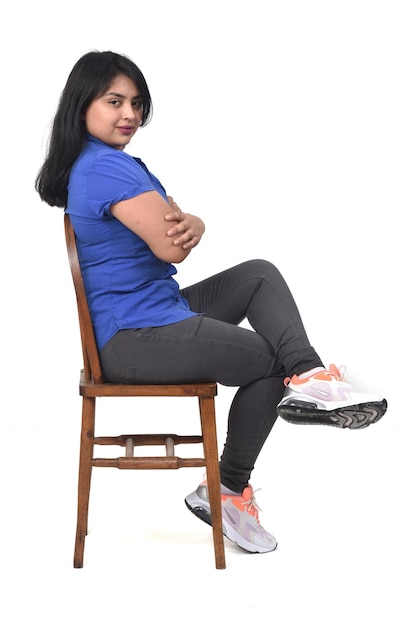 Portrait complet d'une femme latine vêtue de jeans baskets et chemise assise sur charir, bras et jambes croisés sur fond blanc