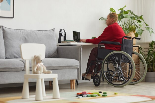 Portrait complet d'une femme métisse moderne utilisant un fauteuil roulant tout en travaillant à domicile au bureau avec des jouets pour enfants au premier plan, espace pour copie