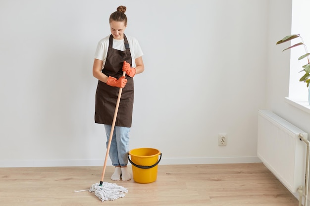 Portrait complet d'une femme portant des gants en caoutchouc orange, un tablier marron et un jean lavant le sol à la maison, regardant en souriant, faisant des tâches ménagères pendant le week-end.