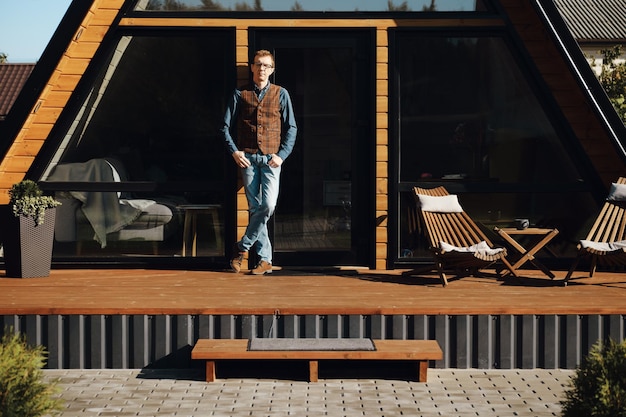 Portrait complet d'un homme âgé en chemise jeans et gilet en laine sur la terrasse d'une cabane en bois