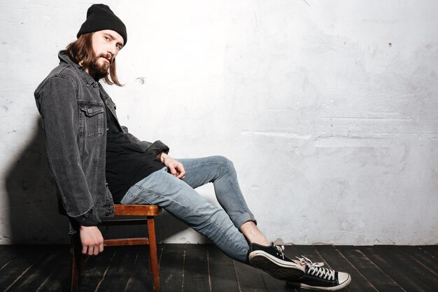 Portrait complet d'un homme barbu élégant au chapeau assis sur une chaise isolée sur le mur