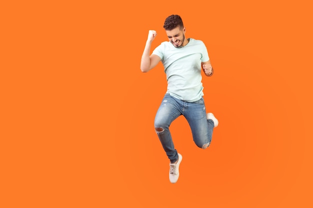 Portrait complet d'un homme brune extatique avec une barbe en baskets et une tenue en jean sautant dans l'air montrant oui je l'ai fait un geste, copiez l'espace pour l'annonce. tourné en studio intérieur isolé sur fond orange