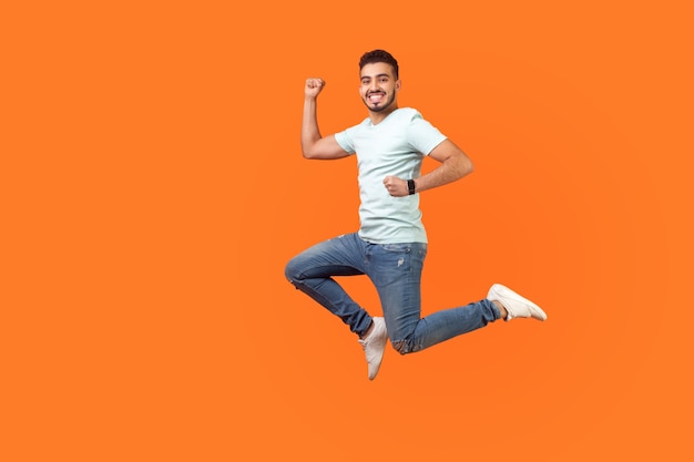 Portrait complet d'un homme brune d'inspiration positive avec une barbe en baskets, une tenue en jean sautant dans l'air ou courant rapidement et rapidement. tourné en studio intérieur isolé sur fond orange, espace de copie vide