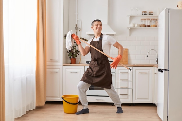 Portrait complet d'un homme de race blanche aux cheveux noirs optimiste positif portant un tablier marron laver le sol dans la cuisine s'amusant tout en nettoyant la maison dansant avec une vadrouille