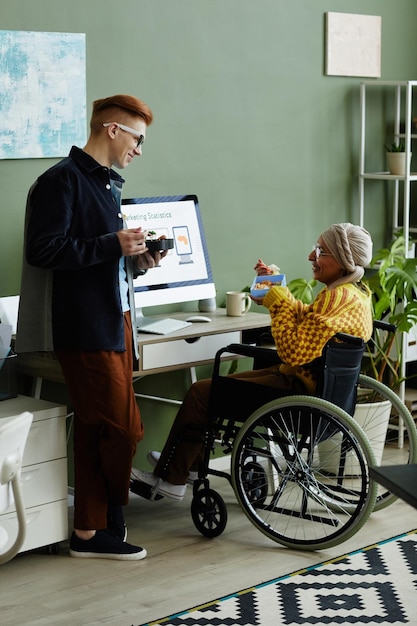 Portrait complet d'une jeune femme utilisant un fauteuil roulant au bureau et déjeunant avec des collègues