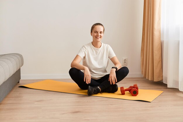 Portrait complet d'une jeune fille sportive assise sur le sol sur un tapis de yoga femme portant des leggins noirs et un t-shirt blanc étant heureuse de terminer son entraînement en regardant la caméra