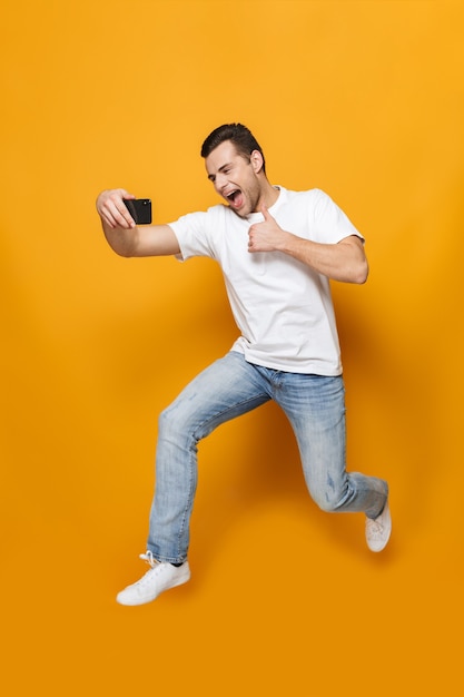 Portrait complet d'un jeune homme heureux portant un t-shirt sautant isolé sur un mur jaune, prenant un selfie