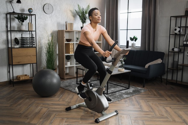 Portrait complet d'une jeune sportive s'entraînant à la salle de sport à domicile faisant du cardio sur un vélo stationnaire