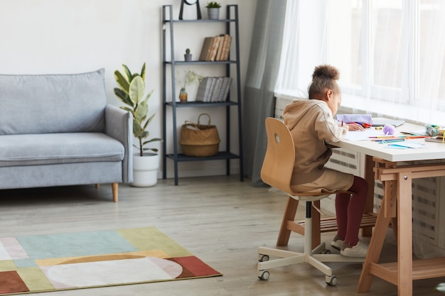 Portrait complet d'une jolie fille afro-américaine faisant ses devoirs ou dessinant assis au bureau dans un intérieur confortable, espace pour copie