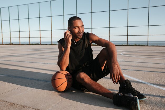 Portrait complet d'un joueur de basket-ball souriant assis sur le terrain de jeu et écoutant de la musique avec des écouteurs