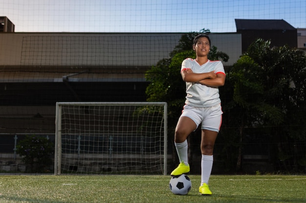 Portrait complet d'une joueuse de football latine avec les bras croisés et un pied sur le ballon sur le terrain de football