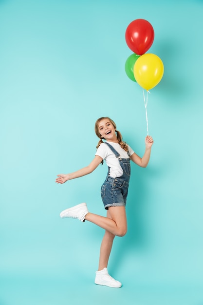 Portrait complet d'une petite fille joyeuse isolée sur un mur bleu, tenant un tas de ballons à air colorés, posant