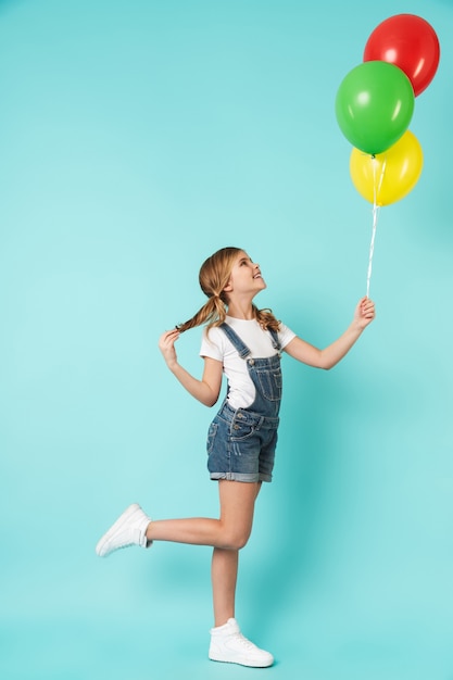 Portrait complet d'une petite fille joyeuse isolée sur un mur bleu, tenant un tas de ballons à air colorés, posant