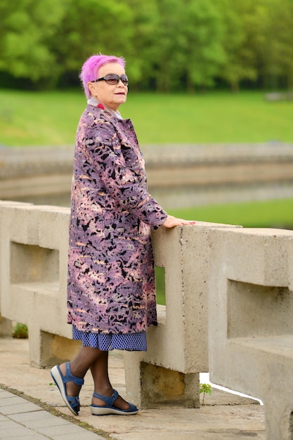 Portrait complet d'une vieille femme aux cheveux roses courts et au manteau de laine debout près du quai de la rivière