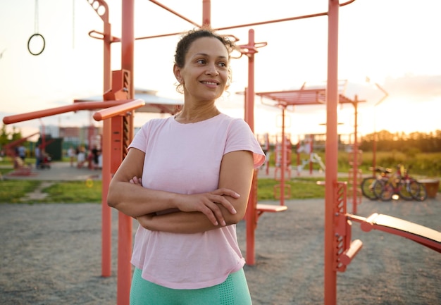 Portrait confiant d'une agréable femme athlétique africaine en vêtements de sport posant avec les bras croisés contre la barre transversale sur le terrain de sport au coucher du soleil