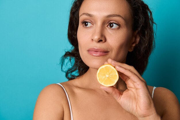 Portrait Confiant En Gros Plan D'une Charmante Femme élégante Et Séduisante De Beauté Naturelle Avec Des Cheveux Noirs Bouclés Isolés Sur Fond Bleu Clair Tenant La Moitié Du Citron. Soins De Santé, Soins De La Peau, Soins Du Corps.