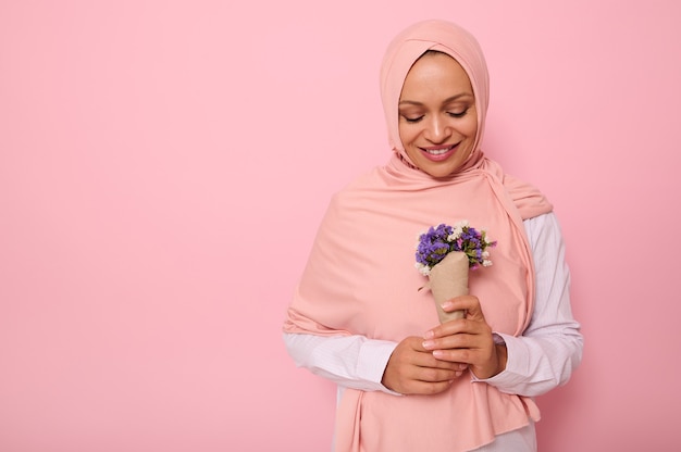 Portrait confiant isolé sur fond coloré avec espace de copie d'une jolie femme arabe musulmane en hijab rose, regardant un joli bouquet de fleurs sauvages dans des tons violets, enveloppé dans du papier kraft