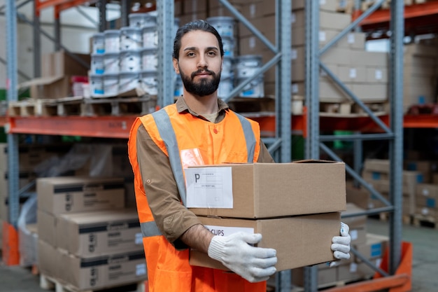 Portrait de contenu beau mixed race mover en gilet réfléchissant transportant des marchandises emballées tout en le préparant pour l'expédition