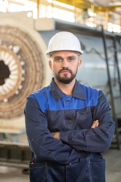 Portrait de contenu brutal homme d'usine barbu dans un casque orange debout avec les bras croisés en atelier