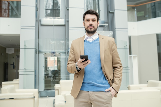 Portrait de contenu jeune avocat barbu en pull bleu tenant la main dans la poche et vérifiant le smartphone dans le hall