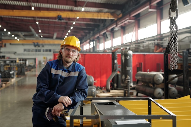 Portrait de contenu jeune ouvrier de production barbu dans un casque jaune et des lunettes de protection s'appuyant sur un cadre métallique à l'usine