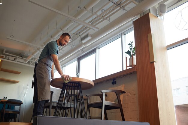 Portrait en contre-plongée d'un serveur masculin nettoyant les tables dans un café ou un café tout en se préparant à l'ouverture le matin, espace de copie