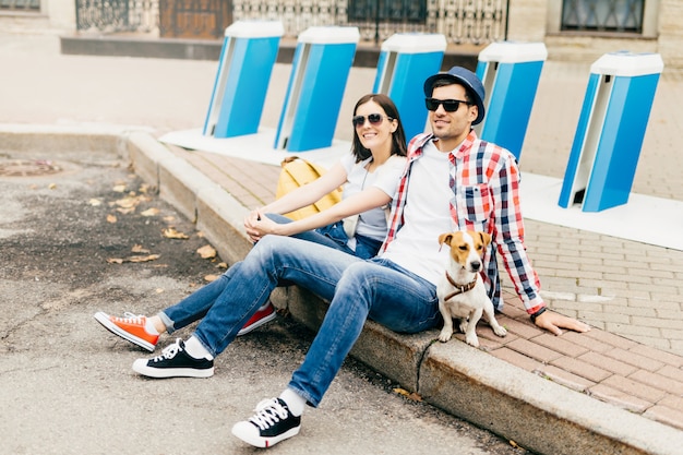 Portrait sur le côté des hommes et des femmes à la mode, portant des lunettes de soleil, assis sur le trottoir à l'extérieur, regardant au loin, pensant où aller, se détendre ensemble. Loisirs, concept de style de vie