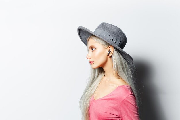 Portrait côté studio d'une jeune jolie fille blonde avec des écouteurs sans fil dans les oreilles portant un chapeau gris et une chemise rose sur fond blanc